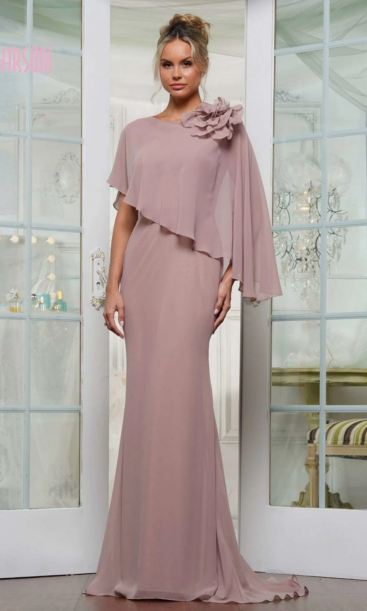 A woman stands indoors in front of glass double doors, wearing the Marsoni MV1347 long formal dress in mauve chiffon, featuring a draped overlay and a large flower detail on one shoulder.